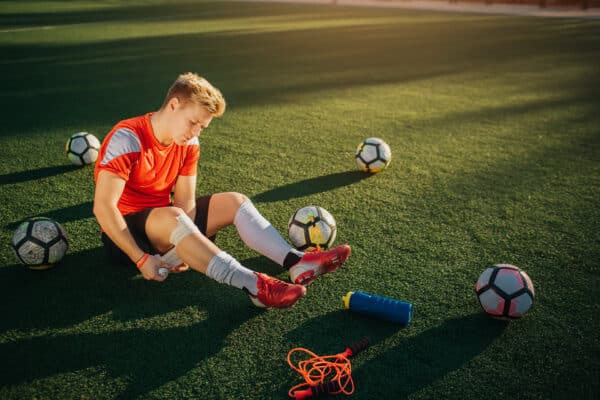 Young player sitting on lawn and wind around leg with bandage. He does it careful. Guy is surround with balls. Theere are bottle of water and jump rope on lawn.